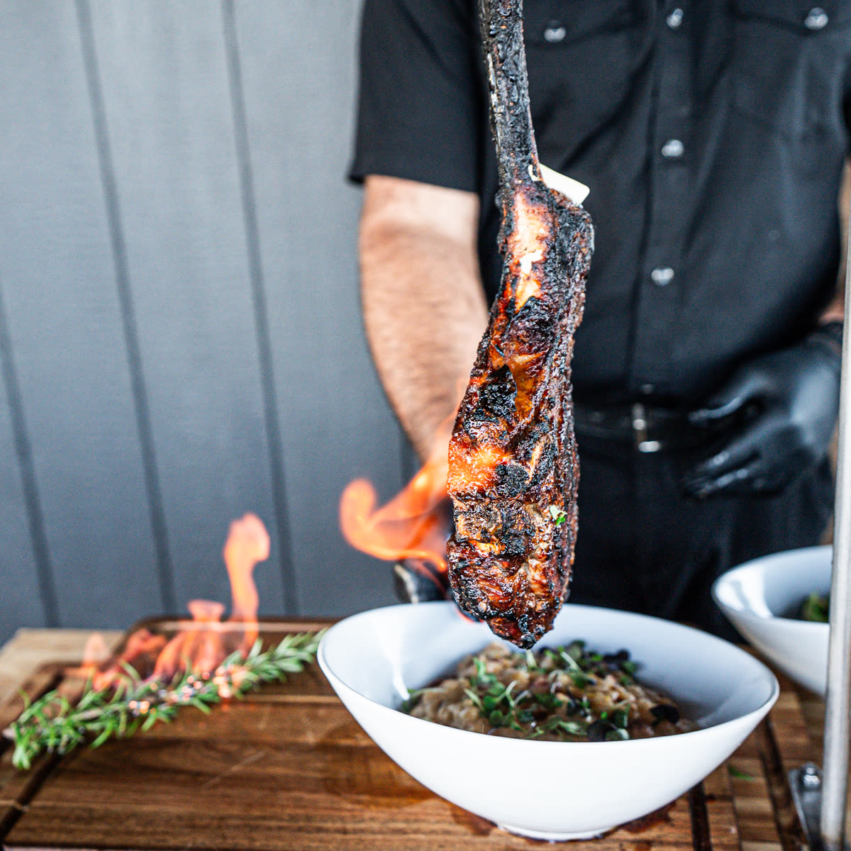 Chef in black gloves plating a flaming tomahawk steak over a bowl, rosemary burning beside it